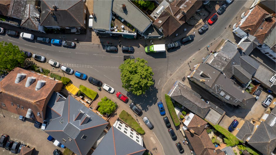 An aerial view of a residential street showing tightly packed houses with tiled and flat roofs, some with chimneys and dormer windows, lining both sides of the road. Numerous parked cars in various colours are situated along the curbs, occupying parallel parking spaces on both sides of the street. A central section of the road curves around a green tree with lush foliage, creating a focal point, while additional vehicles are parked along adjacent side streets and in driveways. The scene is illuminated by natural daylight, casting shadows from the buildings and trees, and demonstrates typical urban house removal logistics with parked vehicles and tight urban street arrangements. Man with Van Gallows Corner would facilitate such home relocations, ensuring furniture transport and packing within this dense, residential environment, often requiring careful loading and unloading during the moving process in the area mentioned in the webpage about avoiding parking fines during moves.