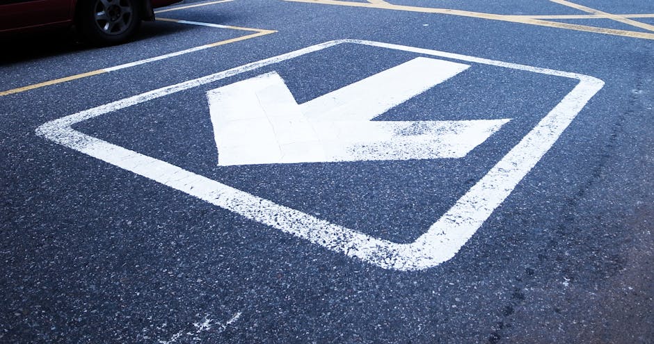 A close-up view of a designated loading zone painted on an asphalt driveway, featuring a white arrow pointing downward within a square outline, indicating an area for vehicle parking or unloading associated with home relocations. The surface around the marking shows some wear and patchy paint, with parking spaces delineated by yellow lines in the background. The scene is outdoors, likely near a property involved in furniture transport or house removals, with natural lighting and no visible vehicles or objects immediately present in the frame. This visual is relevant to strategies for avoiding Havering Council parking fines during a move, as it illustrates a typical loading area that may be used in conjunction with services provided by Man with Van Gallows Corner, a company specialising in removals and relocation logistics.