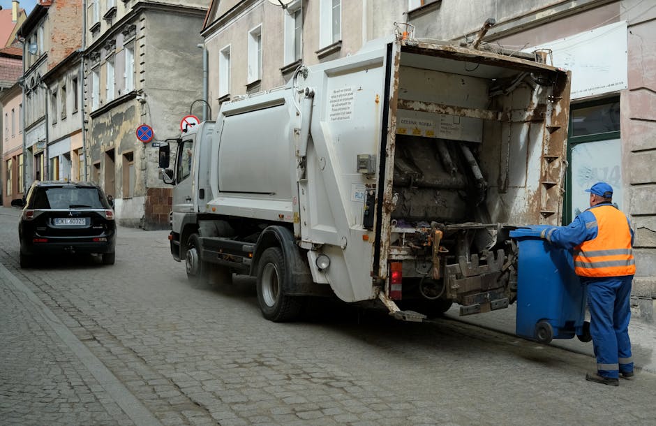 A street scene showing a white waste collection truck parked next to a pavement outside a row of multi-storey residential buildings with mixed brick and stone facades. The rear compactor of the truck is open, revealing rusted metal components and black waste material inside. A worker dressed in blue overalls, a high-visibility orange vest, and a blue cap is placing a blue wheeled waste bin into the truck's open waste intake area. To the left of the truck, a black car is parked on the cobbled street, and there are street signs indicating parking restrictions visible on the building wall. The scene illustrates waste clearance during a home or commercial property waste removal, aligned with professional waste collection services as part of a house removal or clearance process, fitting with the theme of Bulky Waste Removals in Gallows Corner by Man with Van Gallows Corner.