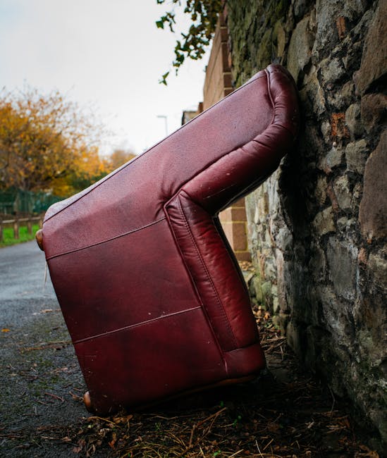 A large, burgundy leather armchair with visible wear and creases, positioned outdoors on a paved path next to a stone wall. The armchair is resting at an angle, with the backrest leaning against the wall and the base on the ground. Surrounding the chair are fallen leaves and some dirt, indicating an outdoor setting. In the background, there are trees with autumn foliage and a cloudy sky, suggesting it is autumn. The scene appears to involve a home relocation or furniture transport process, possibly part of a house move arranged by Man with Van Gallows Corner, with the furniture being prepared for loading or unloading during a removal service.