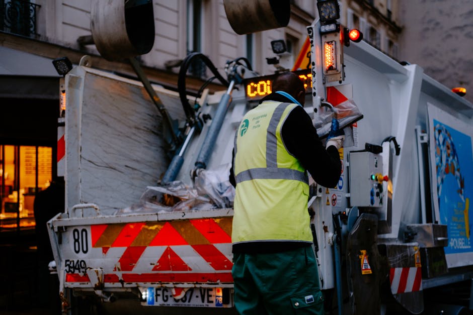 A street scene showing a white waste collection truck parked next to a pavement outside a row of multi-storey residential buildings with mixed brick and stone facades. The rear compactor of the truck is open, revealing rusted metal components and black waste material inside. A worker dressed in blue overalls, a high-visibility orange vest, and a blue cap is placing a blue wheeled waste bin into the truck's open waste intake area. To the left of the truck, a black car is parked on the cobbled street, and there are street signs indicating parking restrictions visible on the building wall. The scene illustrates waste clearance during a home or commercial property waste removal, aligned with professional waste collection services as part of a house removal or clearance process, fitting with the theme of Bulky Waste Removals in Gallows Corner by Man with Van Gallows Corner.