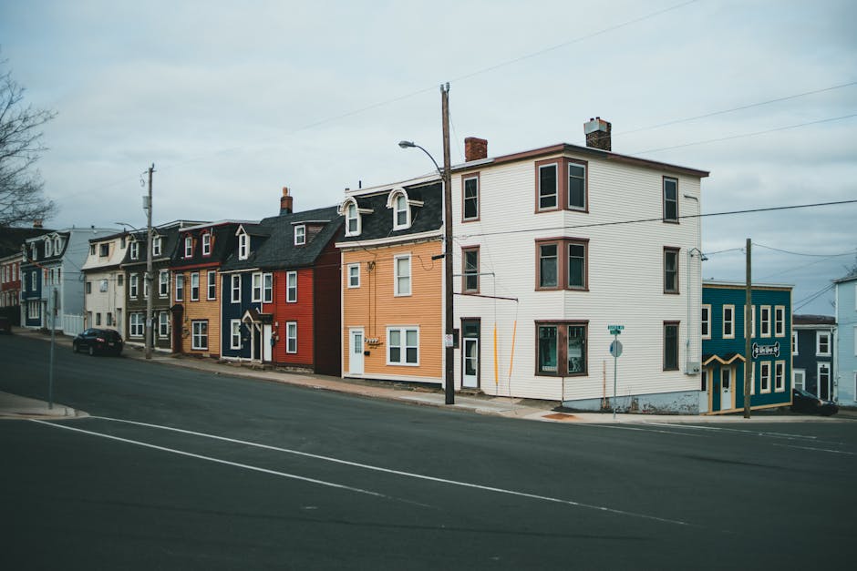 A row of three-storey residential buildings along a quiet street, featuring a variety of exterior colours including white, red, yellow, and blue. The buildings have multiple windows, some with bay window sections, and the street is empty with no visible pedestrians. A black car is parked on the side of the road near the houses. Utility poles and wires run along the street, and a street lamp is positioned near the corner of the white building. In the foreground, a section of the paved road is visible with white road markings. The scene appears to be during daytime with overcast sky, and the environment suggests a typical neighbourhood setting suitable for house removals and moving services, such as those provided by Man with Van Gallows Corner, supporting logistics like packing, furniture transport, and loading processes.