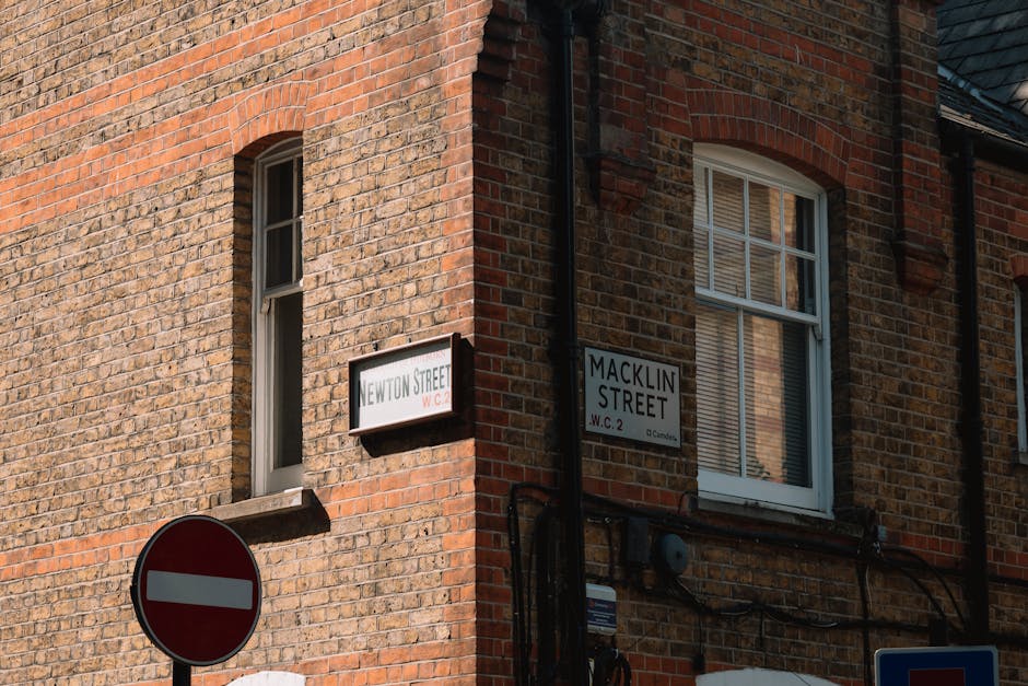 A close-up view of the corner of a brick building at the intersection of Newton Street and Macklin Street, with street signs visible on the wall. The building has a red and brown brick exterior, with two white-framed windows, one of which is partly open with blinds inside. A black downspout runs vertically along the corner, and several black cables are attached to the wall below the signs. In the lower left corner, a 'no entry' traffic sign with a red circle and white horizontal bar is partially visible. The scene is illuminated by natural daylight, highlighting the textures of the brickwork and the signage, which are relevant to logistics and house removals in the area, as well as moving permit considerations for Gallows Corner.