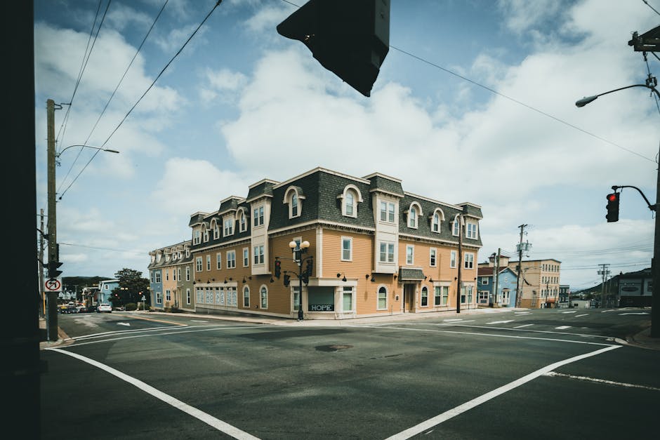 A row of three-storey residential buildings along a quiet street, featuring a variety of exterior colours including white, red, yellow, and blue. The buildings have multiple windows, some with bay window sections, and the street is empty with no visible pedestrians. A black car is parked on the side of the road near the houses. Utility poles and wires run along the street, and a street lamp is positioned near the corner of the white building. In the foreground, a section of the paved road is visible with white road markings. The scene appears to be during daytime with overcast sky, and the environment suggests a typical neighbourhood setting suitable for house removals and moving services, such as those provided by Man with Van Gallows Corner, supporting logistics like packing, furniture transport, and loading processes.
