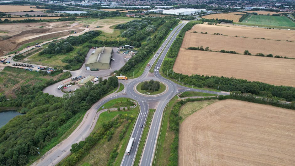 An aerial view of the Gallows Corner roundabout showing multiple lanes of traffic including trucks and cars moving around the circular junction surrounded by agricultural fields, patches of greenery, and a few industrial buildings. The roundabout connects the A127 road with a smaller road leading to nearby properties. The landscape features open farmland with large rectangular fields, some with planted crops and others with bare soil, interspersed with line hedges and trees. The scene captures the infrastructure of busy home relocation and furniture transport routes, consistent with services offered by Man with Van Gallows Corner for house removals and moving logistics, with the roundabout facilitating efficient route planning for home relocations across the area.