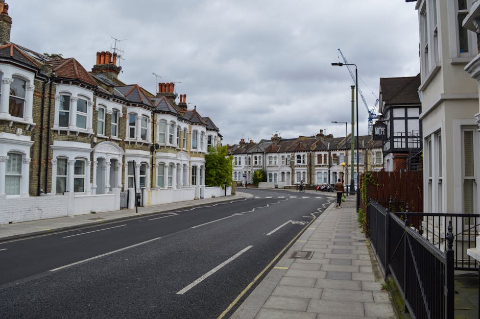 A quiet residential street scene during daytime with a paved road lined by multi-story brick and stone houses, some with small front gardens enclosed by low brick walls and lush green hedges. Several 