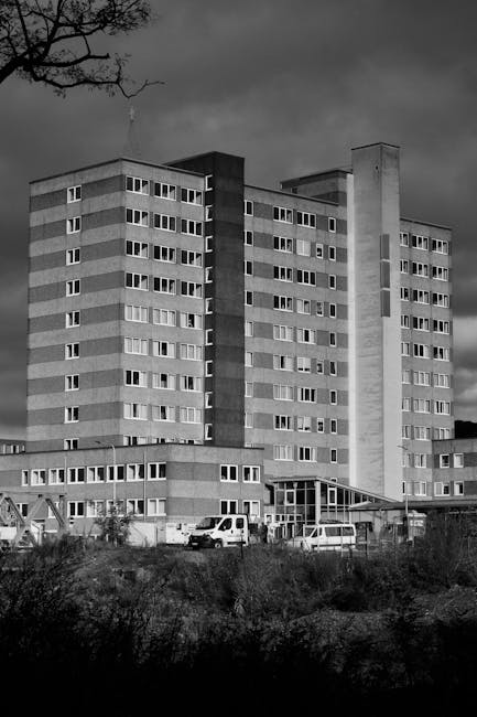 A black and white photograph of a large, multi-storey residential apartment block with multiple rows of windows. The building features a flat roof and a prominent central vertical section in a darker shade, contrasting with the lighter exterior walls. In the foreground, there is a construction or loading area with several parked vehicles, including a white van and a few cars, situated on an uneven patch of land with visible dirt and sparse vegetation. The environment appears under cloudy skies, suggesting overcast weather. Part of a leafless tree is visible at the top left corner of the image. Inside the property, near the base of the building, there are moving or loading activities occurring, possibly involving packaging materials and equipment, which aligns with the house removals and moving context often related to furniture transport and packing processes. The scene is associated with relocation services by Man with Van Gallows Corner, providing insights into the logistics of home moves.