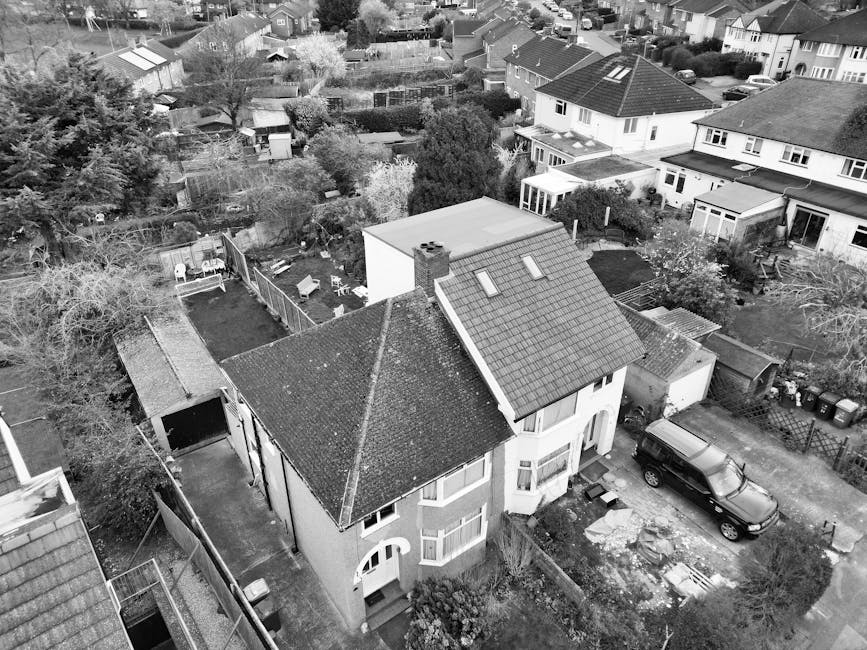 An aerial black-and-white photograph of a residential neighbourhood showing a two-storey house with a pitched tiled roof, situated on a corner plot, surrounded by neighboring houses, gardens, and tree