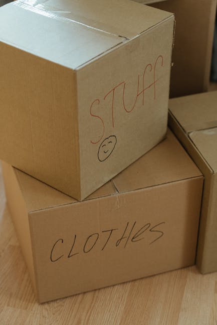 A close-up of three cardboard moving boxes placed on a wooden floor inside a home, prepared for housing items during a house relocation. The top box has the word 'STUFF' written in red marker, accompanied by a small smiley face, while the bottom box is labeled 'CLOTHES' in black marker. The boxes are sealed with packing tape to secure contents. The background shows a partially visible cardboard box, indicating an organized packing and moving process. These boxes are ready for transportation as part of a home removal service, highlighting packing preparations for furniture transport and belongings transfer between properties. The image suggests a professional move by Man with Van Gallows Corner, focusing on efficient packing and loading during local removals.