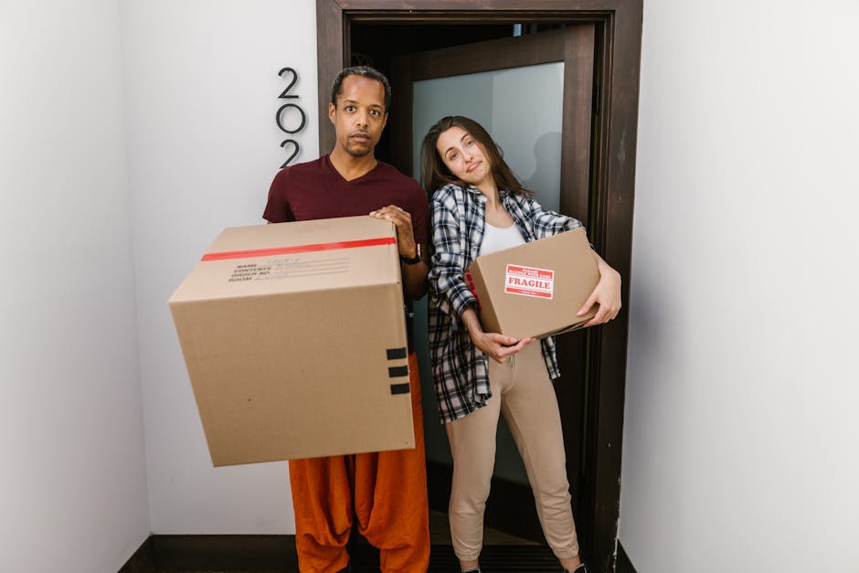 A man and a woman standing inside a doorway of a residential property, each holding a cardboard moving box. The man appears to be lifting a large box with black tape seals and a red stripe, while the woman holds a smaller, labeled fragile box. Both individuals look directly at the camera with neutral expressions. The background shows a white wall with the house number partially visible and a dark wooden doorframe. The scene is illuminated by indoor lighting, suggesting an indoor packing and home relocation process. The setting indicates a typical room prepared for a house move, with the boxes being part of the packing and furniture transport carried out by Man with Van Gallows Corner, a professional removals service experienced in local moves within Harold Hill and Romford.