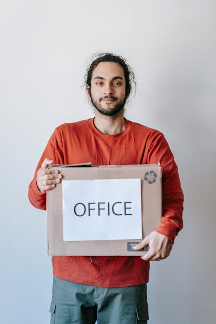 A man in formal office attire, holding a cardboard box, is standing in a modern office environment with a grey wall background. The desk in front of him has several white binders with labels, a black