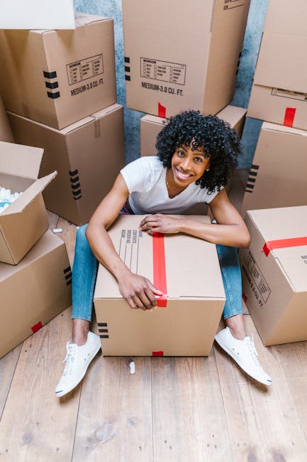 A smiling woman with curly black hair, dressed in a white t-shirt and blue jeans, sitting on a wooden floor surrounded by numerous large cardboard boxes sealed with red packing tape. The boxes are of various sizes, some stacked against a blue wall and others on the floor, with open flaps revealing packing materials inside. The woman is holding a box on her lap with one hand, while her other arm rests on another box beside her. The scene depicts the packing phase of a home relocation, with the boxes prepared for transport by [COMPANY_NAME]. The lighting is bright and natural, emphasizing the organized and detailed packing environment typical of professional furniture transport and moving services. This image illustrates the process of packing and moving within a house, aligned with the services offered under removals near Gallows Corner, as referenced in the page about the best times to move in Gallows Corner.