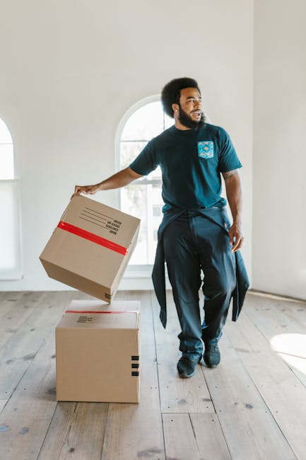 A man with dark curly hair and a beard, dressed in a dark blue t-shirt and matching trousers, is inside a bright, empty room with large arched windows allowing natural light to fill the space. He is holding a medium-sized cardboard box sealed with red tape in his right hand, while standing next to two more cardboard boxes placed on a light wood floor. The boxes appear to be part of packing materials used for home relocation, and the room has plain white walls with minimal furnishings. This scene exemplifies a typical furniture transport and packing process involved in a professional house removal, with a focus on careful handling and preparation for moving services provided by Man with Van Gallows Corner, as part of their removals expertise.