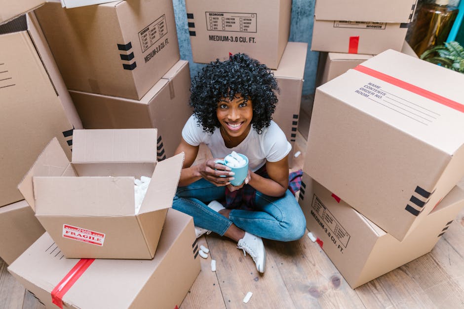 A smiling woman with curly black hair, dressed in a white t-shirt and blue jeans, sitting on a wooden floor surrounded by numerous large cardboard boxes sealed with red packing tape. The boxes are of various sizes, some stacked against a blue wall and others on the floor, with open flaps revealing packing materials inside. The woman is holding a box on her lap with one hand, while her other arm rests on another box beside her. The scene depicts the packing phase of a home relocation, with the boxes prepared for transport by [COMPANY_NAME]. The lighting is bright and natural, emphasizing the organized and detailed packing environment typical of professional furniture transport and moving services. This image illustrates the process of packing and moving within a house, aligned with the services offered under removals near Gallows Corner, as referenced in the page about the best times to move in Gallows Corner.