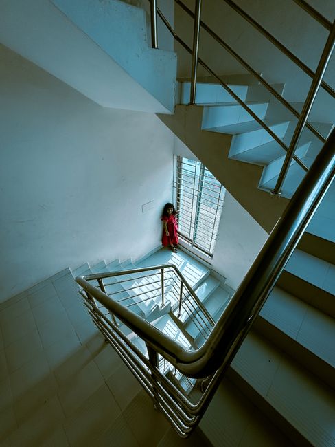A multi-level staircase inside a building with white tiled steps and a stainless steel handrail. The staircase curves around a central light gray wall, with a large glass window at the landings allowing natural light to illuminate the area. A small doll dressed in pink with dark hair, positioned near the window, is visible at the base of the staircase. The environment appears clean and modern, with an open, spacious design typical of residential or commercial stairwells, aligned with house relocations and furniture transport processes managed by Man with Van Gallows Corner.