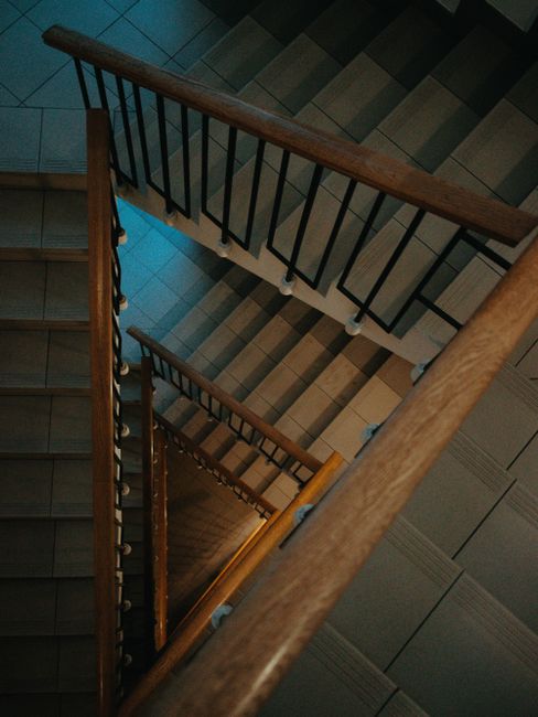 A view looking directly down a wooden spiral staircase with black metal balustrades, situated inside a building with tiled flooring in shades of brown and gray. The staircase is actively being used in the context of a house removal, with a silver metal trolley visible on the lower steps, carrying cardboard boxes wrapped in plastic and packing materials. The scene captures part of the home interior, including the stairwell's wooden handrail and tiled floor, highlighting the logistical aspect of furniture transport during a relocation managed by Man with Van Gallows Corner. The lighting is natural, and the image emphasizes the vertical movement involved in packing and moving household items during a house relocation process.