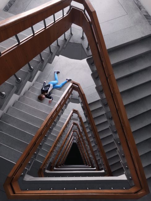 A multi-level staircase inside a building with white tiled steps and a stainless steel handrail. The staircase curves around a central light gray wall, with a large glass window at the landings allowing natural light to illuminate the area. A small doll dressed in pink with dark hair, positioned near the window, is visible at the base of the staircase. The environment appears clean and modern, with an open, spacious design typical of residential or commercial stairwells, aligned with house relocations and furniture transport processes managed by Man with Van Gallows Corner.