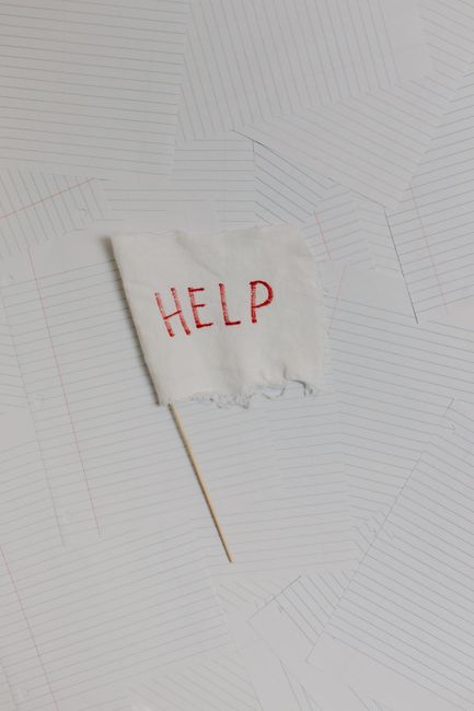 A small white fabric flag with the word 'HELP' embroidered in red thread is attached to a thin, light wooden stick. The flag lies flat on a surface covered with architectural or site plans, which display detailed, overlapping line drawings and patterns in light grey and subtle red. The surface appears to be a paper plan or blueprint, likely used for a property or building project. There are no additional objects or furniture visible, and the lighting is neutral, evenly illuminating the scene. This image illustrates a conceptual call for assistance, potentially related to urgent eviction or home relocation situations, aligning with the theme of removals and moving services provided by Man with Van Gallows Corner.