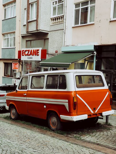 A vintage orange and white van parked on a cobblestone street in front of a multistory residential building with pastel-colored facades and multiple windows. The van has a retro design with a large rear door, small windows along the side, and is positioned close to the pavement. Behind the van, a green awning extends over a storefront, and a sign reading 'ECZANE' (pharmacy) is visible on the building. The setting suggests a typical urban area where home relocation or furniture transport services, such as those provided by Man with Van Gallows Corner, might operate. The image captures a loading or unloading scenario, with the van's positioning implying a moving process where access might be limited, highlighting the importance of careful vehicle placement during house removals or packing and moving tasks.