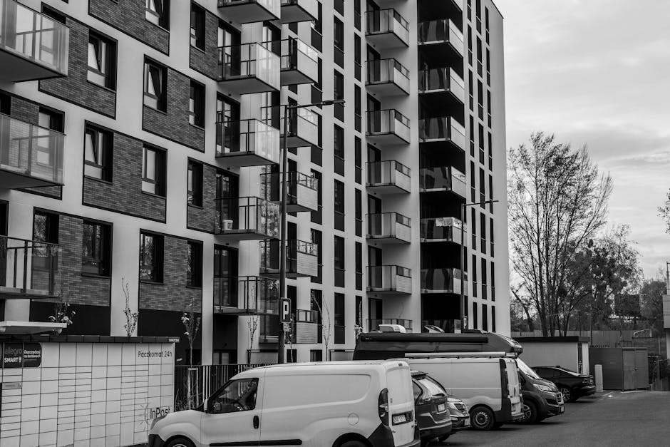 A black and white photograph of a modern, multi-storey residential building with multiple balconies, situated in Gallows Corner. In the foreground, several parked vehicles, including a white van, a cargo van, and a smaller car, are positioned along the street outside the building. The building's ground floor features large windows and a vehicle entrance, with a parking sign visible near the entrance. The street area appears as a designated loading zone or parking area for home relocation and furniture transport services. Surrounding the building are a few leafless trees, and the sky appears overcast. This scene exemplifies typical logistics challenges faced when a van cannot park directly outside a flat, necessitating careful loading and moving procedures that [COMPANY_NAME] would manage as part of their removals service.