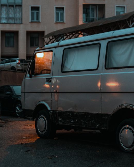 A vintage orange and white van parked on a cobblestone street in front of a multistory residential building with pastel-colored facades and multiple windows. The van has a retro design with a large rear door, small windows along the side, and is positioned close to the pavement. Behind the van, a green awning extends over a storefront, and a sign reading 'ECZANE' (pharmacy) is visible on the building. The setting suggests a typical urban area where home relocation or furniture transport services, such as those provided by Man with Van Gallows Corner, might operate. The image captures a loading or unloading scenario, with the van's positioning implying a moving process where access might be limited, highlighting the importance of careful vehicle placement during house removals or packing and moving tasks.